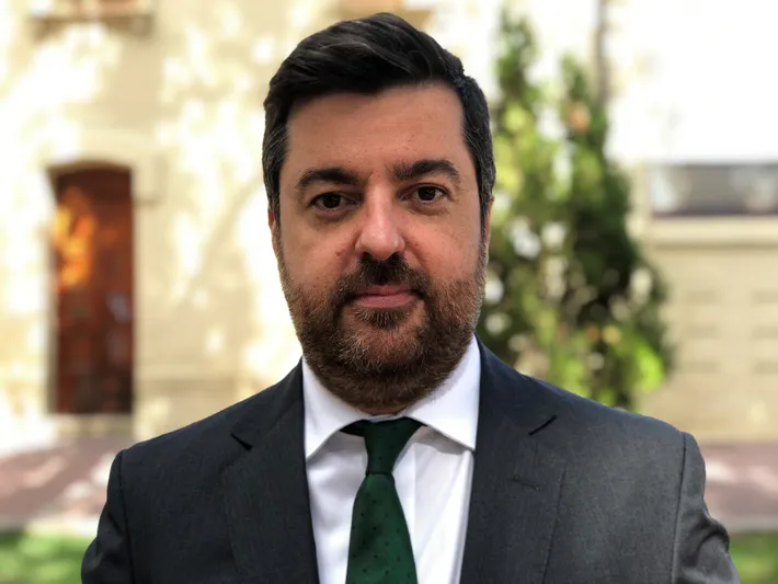 Hombre con pelo y barba morena de frente con americana negra, camisa blanca y corbata verde
