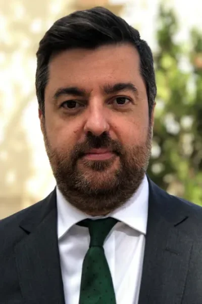 Hombre con pelo y barba morena de frente con americana negra, camisa blanca y corbata verde
